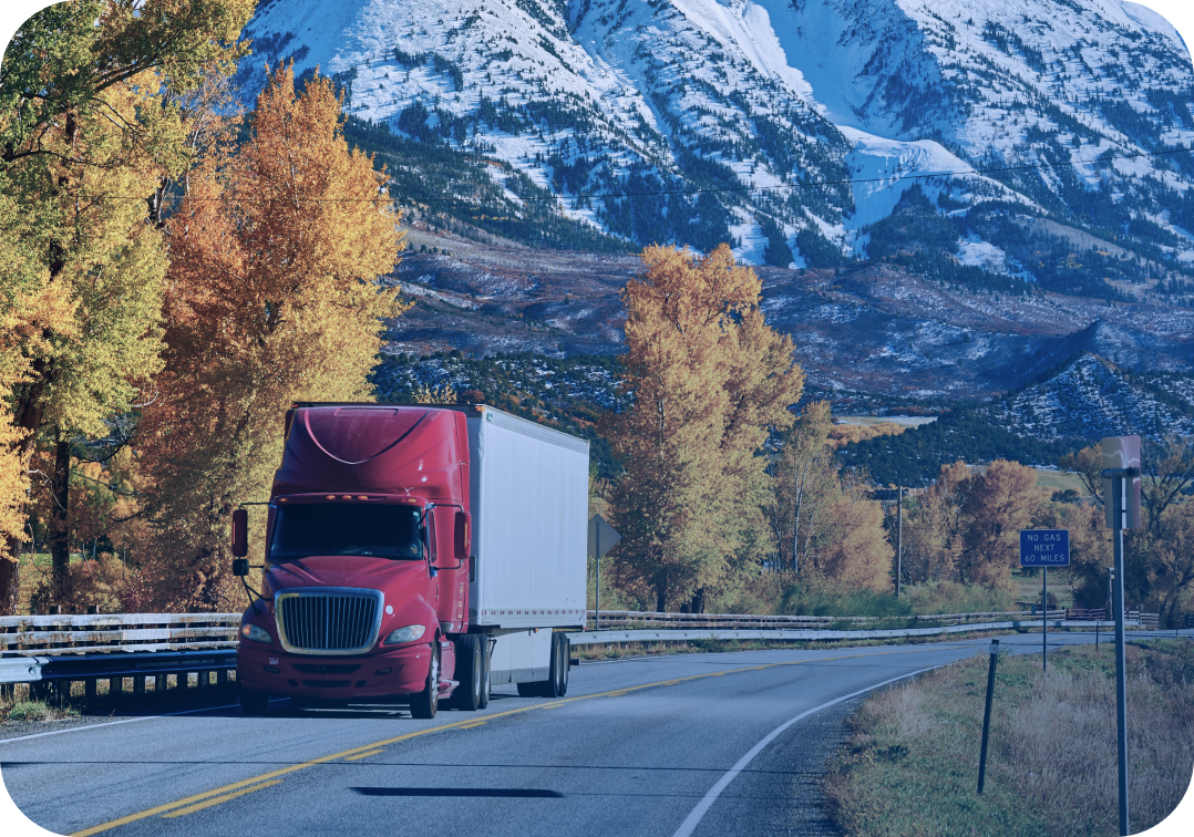 Red truck driving on a scenic mountain highway during autumn, with colorful foliage and snowy peaks in the background.