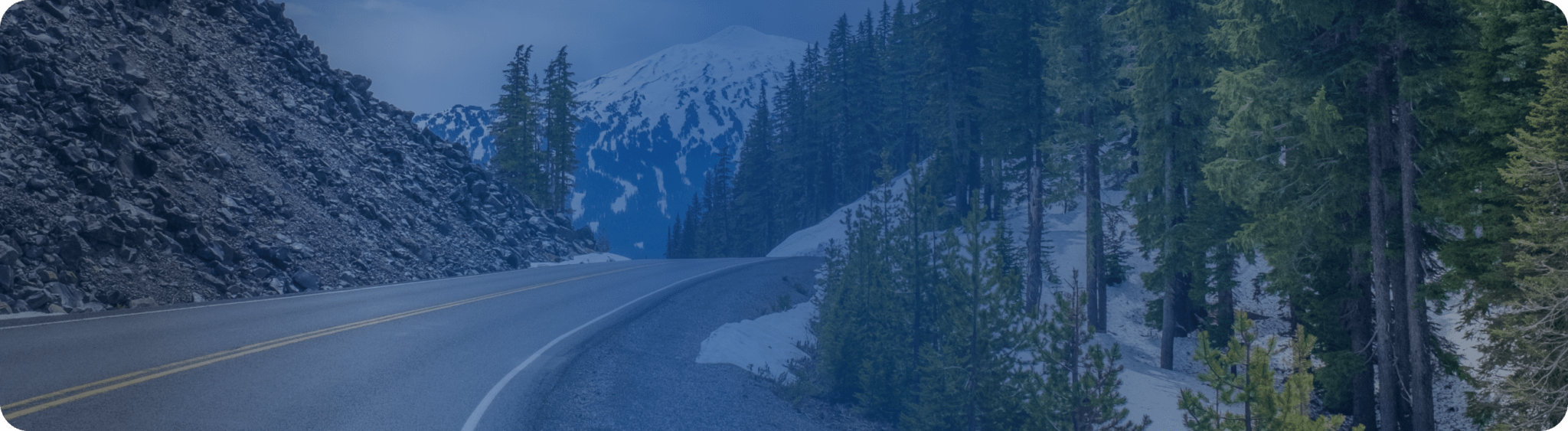 Snow-lined mountain road winding through a dense forest of tall evergreen trees, with a snowy peak visible in the background