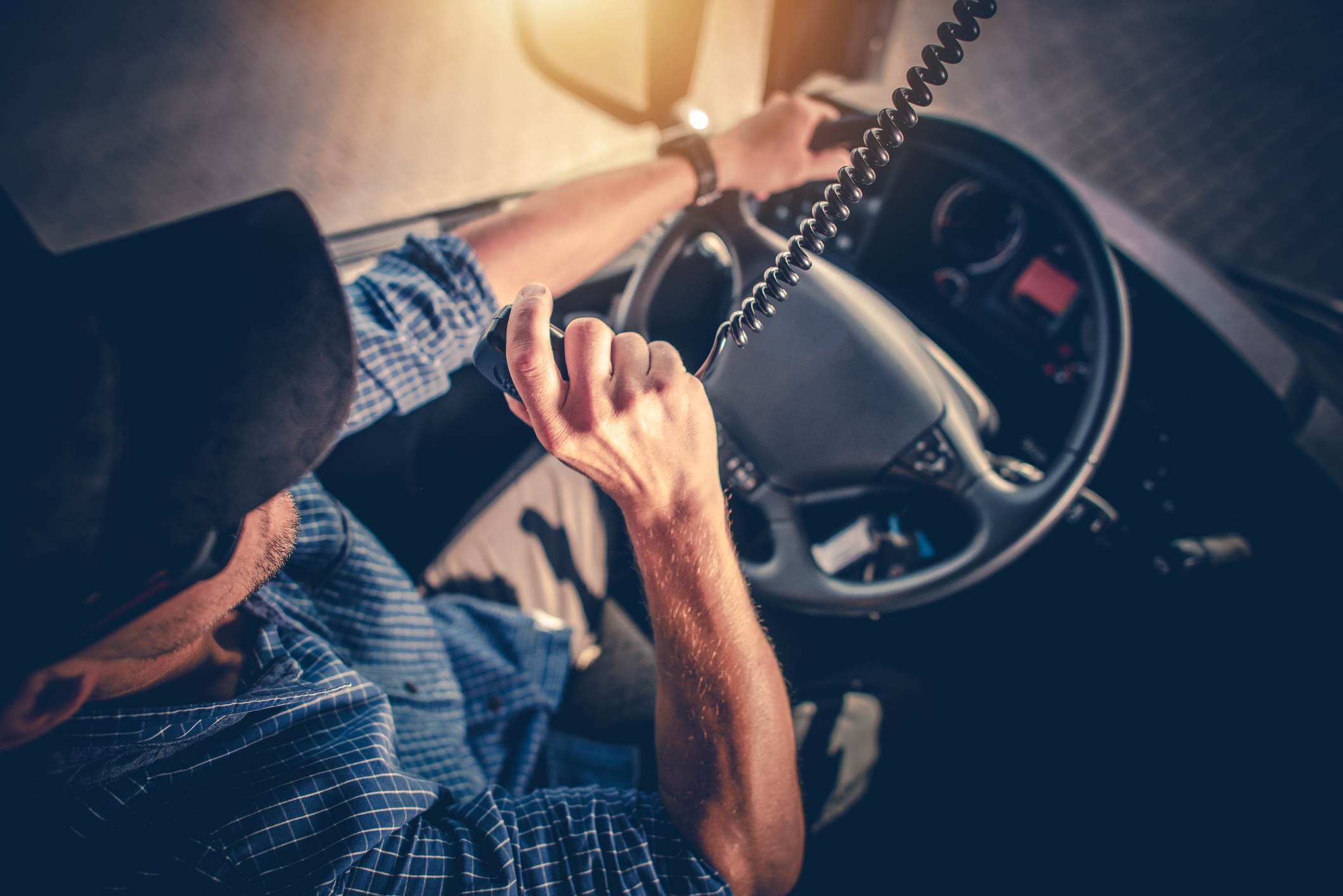 A truck driver in a cap and blue plaid shirt communicates using a CB radio inside the truck cab.